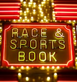 Bright neon lights on a marquee sign display the words 'RACE & SPORTS BOOK'. The sign is surrounded by rows of yellow and white lights against a red background, evoking a vibrant, energetic atmosphere typically associated with casinos or entertainment venues.