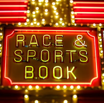 Bright neon lights on a marquee sign display the words 'RACE & SPORTS BOOK'. The sign is surrounded by rows of yellow and white lights against a red background, evoking a vibrant, energetic atmosphere typically associated with casinos or entertainment venues.
