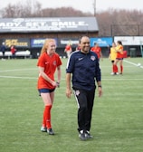 A group of people are on a soccer field, with some wearing sports attire. In the foreground, a person in red sports clothing is walking alongside someone in a dark tracksuit. Other people are visible in the background, some wearing yellow jerseys. A building with advertising banners, including a prominent sign reading 'Ready, Set, Smile,' is in the background.