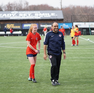 A group of people are on a soccer field, with some wearing sports attire. In the foreground, a person in red sports clothing is walking alongside someone in a dark tracksuit. Other people are visible in the background, some wearing yellow jerseys. A building with advertising banners, including a prominent sign reading 'Ready, Set, Smile,' is in the background.