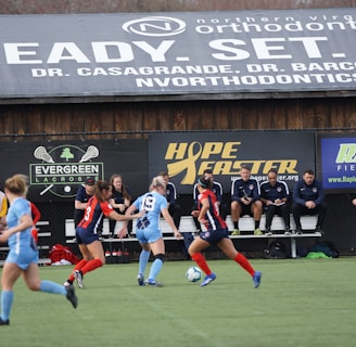 Players engage in a competitive soccer game on a field while others are seated on the bench watching attentively. The action is taking place in front of a wooden structure displaying multiple advertisements, including orthodontics, lacrosse, and field hockey.