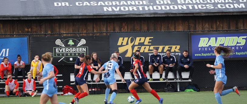 Players engage in a competitive soccer game on a field while others are seated on the bench watching attentively. The action is taking place in front of a wooden structure displaying multiple advertisements, including orthodontics, lacrosse, and field hockey.