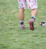 A person wearing sports shoes and patterned shorts stands on a grassy field, with various sports equipment like gloves and a helmet placed on the ground nearby.