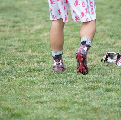 A person wearing sports shoes and patterned shorts stands on a grassy field, with various sports equipment like gloves and a helmet placed on the ground nearby.