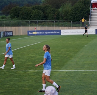 A soccer game scene on a green grass field with players wearing light blue jerseys and white shorts. A goalpost is visible in the background, and there are people sitting or standing on the bleachers. Advertising banners are displayed along the sidelines.