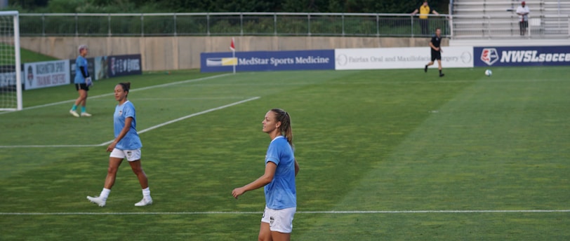 A soccer game scene on a green grass field with players wearing light blue jerseys and white shorts. A goalpost is visible in the background, and there are people sitting or standing on the bleachers. Advertising banners are displayed along the sidelines.