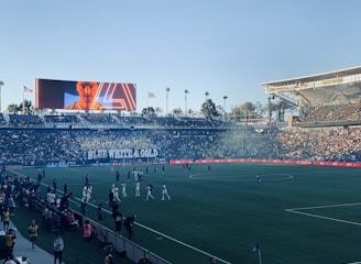 A large sports stadium filled with spectators. The field is occupied by soccer players, some walking and others scattered across the pitch. A massive screen displays a person, and flags fly on top of the stadium roof. The audience is dense, and the atmosphere seems lively with a visible banner showing 'BLUE WHITE & GOLD'.
