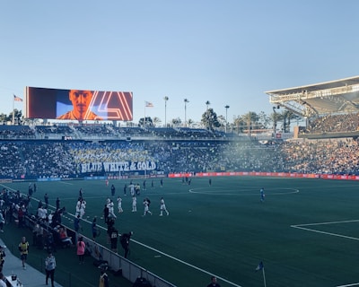 A large sports stadium filled with spectators. The field is occupied by soccer players, some walking and others scattered across the pitch. A massive screen displays a person, and flags fly on top of the stadium roof. The audience is dense, and the atmosphere seems lively with a visible banner showing 'BLUE WHITE & GOLD'.