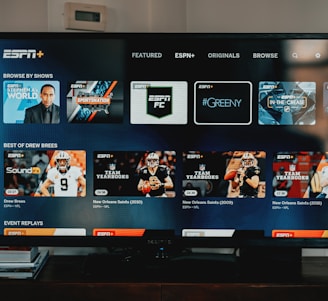 A television screen displaying the ESPN+ streaming interface with various sports-related shows and categories. The screen includes sections such as 'Browse by Shows' and 'Best of Drew Brees' with thumbnails of sports programs featuring athletes in action and analysts.