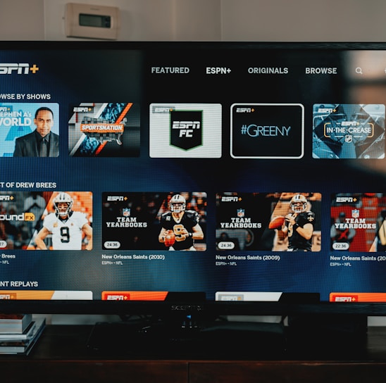 A television screen displaying the ESPN+ streaming interface with various sports-related shows and categories. The screen includes sections such as 'Browse by Shows' and 'Best of Drew Brees' with thumbnails of sports programs featuring athletes in action and analysts.