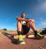 A person wearing a black athletic outfit is seated on a wooden platform in an outdoor sports field. The individual is relaxed, with arms resting on knees. The field is expansive with a clear blue sky and scattered clouds above. A brightly colored running shoe and athletic gear are visible, along with a water bottle and a blue backpack placed beside the person. Lampposts and residential buildings can be seen in the background.