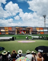 A sports stadium with a large stage set up on the field, hosting an outdoor event. Spectators wearing colorful hats sit in the foreground. The seating areas display vibrant colors, and a clear blue sky with scattered clouds is visible. Large screens display graphics related to the event.