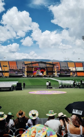 A sports stadium with a large stage set up on the field, hosting an outdoor event. Spectators wearing colorful hats sit in the foreground. The seating areas display vibrant colors, and a clear blue sky with scattered clouds is visible. Large screens display graphics related to the event.
