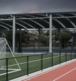 An outdoor sports facility featuring a running track and a soccer goal. The track is red, surrounded by a black metal fence, and the field is covered with artificial green turf. In the background, a basketball court is visible under a covered roof with metal supports. The sky is overcast, adding a moody atmosphere.