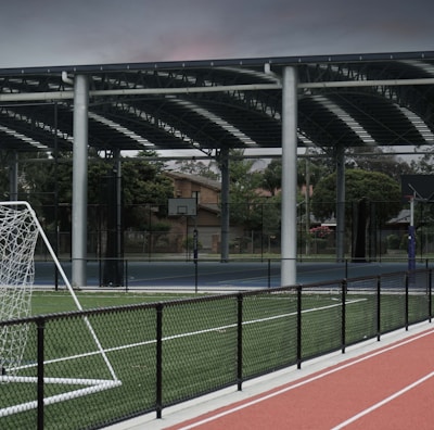 An outdoor sports facility featuring a running track and a soccer goal. The track is red, surrounded by a black metal fence, and the field is covered with artificial green turf. In the background, a basketball court is visible under a covered roof with metal supports. The sky is overcast, adding a moody atmosphere.
