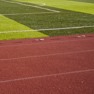 A sports field with vivid green grass divided into sections by white lines in the background. In the foreground, a track with a reddish-brown surface is marked with numbers and lines.