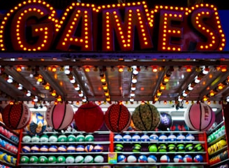 A brightly lit carnival game stall with a large 'GAMES' sign at the top surrounded by red and yellow lights. Various sports balls, including basketballs and soccer balls, are hanging from the ceiling and neatly arranged on shelves below.