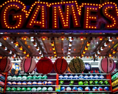 A brightly lit carnival game stall with a large 'GAMES' sign at the top surrounded by red and yellow lights. Various sports balls, including basketballs and soccer balls, are hanging from the ceiling and neatly arranged on shelves below.