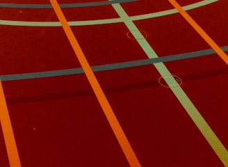 A sports court with intersecting lines in orange and blue, creating geometric patterns on a reddish-brown surface.