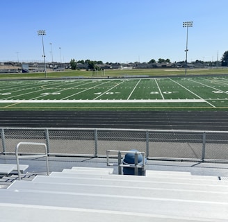 A sports field with lines marking football or soccer measurements is surrounded by a running track. Several tall floodlights are positioned around the area and empty metal bleachers can be seen in the foreground. In the distance, there are trees and buildings under a clear blue sky.