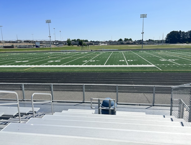 A sports field with lines marking football or soccer measurements is surrounded by a running track. Several tall floodlights are positioned around the area and empty metal bleachers can be seen in the foreground. In the distance, there are trees and buildings under a clear blue sky.