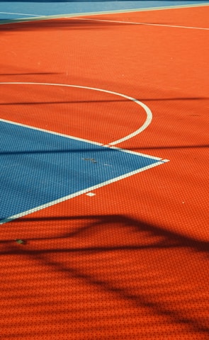 A vibrant sports court with sections of red and blue, featuring crisp white lines marking the boundaries. Shadows from a structure create geometric patterns across the surface, adding depth and interest to the scene.