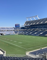 A large, empty sports stadium with blue seats surrounding a well-maintained green grass field. Bright sunlight creates clear shadows, and there are two goal posts on opposite ends of the field. A big electronic scoreboard displays some graphics, and the sky is clear and blue.