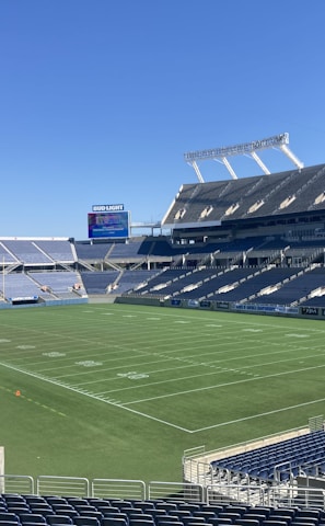 A large, empty sports stadium with blue seats surrounding a well-maintained green grass field. Bright sunlight creates clear shadows, and there are two goal posts on opposite ends of the field. A big electronic scoreboard displays some graphics, and the sky is clear and blue.