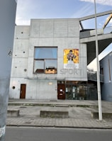 A modern concrete building with a minimalistic design, featuring a large window on the second floor and a poster of a boxer on the exterior wall. The word 'SPORT' is embedded in the concrete wall. The building entrance has glass doors and a bicycle rack is visible on the sidewalk. A streetlight and street are in the foreground, with a clear sky above.