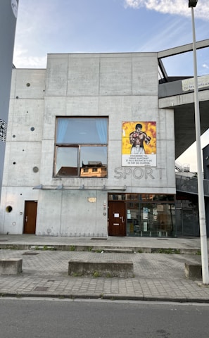 A modern concrete building with a minimalistic design, featuring a large window on the second floor and a poster of a boxer on the exterior wall. The word 'SPORT' is embedded in the concrete wall. The building entrance has glass doors and a bicycle rack is visible on the sidewalk. A streetlight and street are in the foreground, with a clear sky above.
